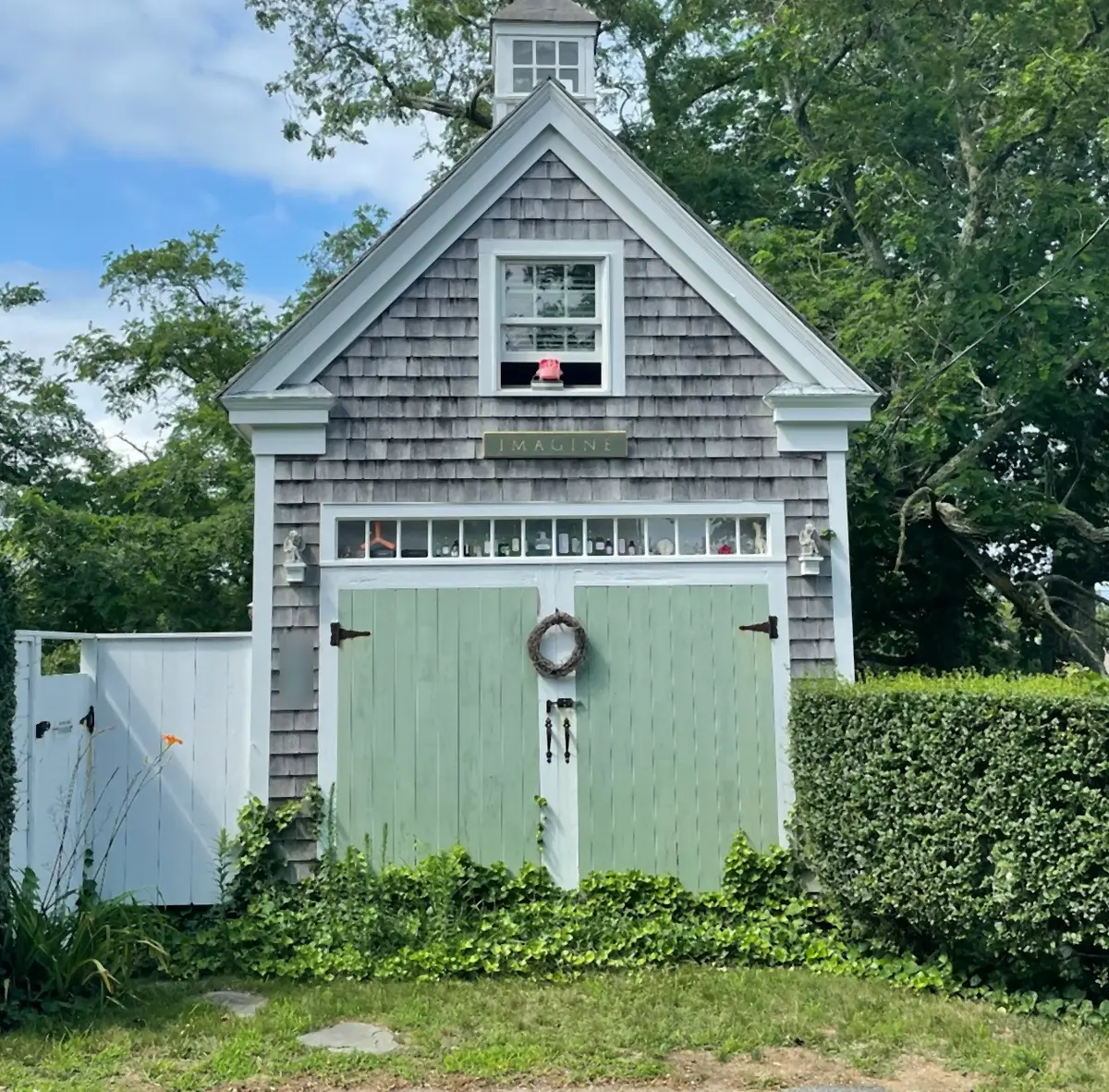 A cedar-shingled Cape Cod carriage house with sage green doors, a wreath and a cupola, between clipped hedges